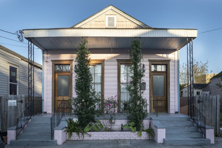Pink shotgun house with two wooden doors, large windows, and climbing plants on the porch.
