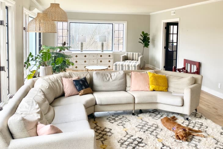 Spacious living room with a beige sectional sofa, colorful pillows, woven pendant lights, plants, and a dog on a patterned rug.