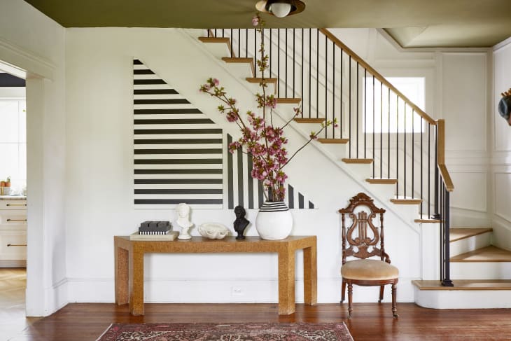 Entryway with a wooden console table, decorative busts, a vase with pink flowers, and a striped wall under a staircase.