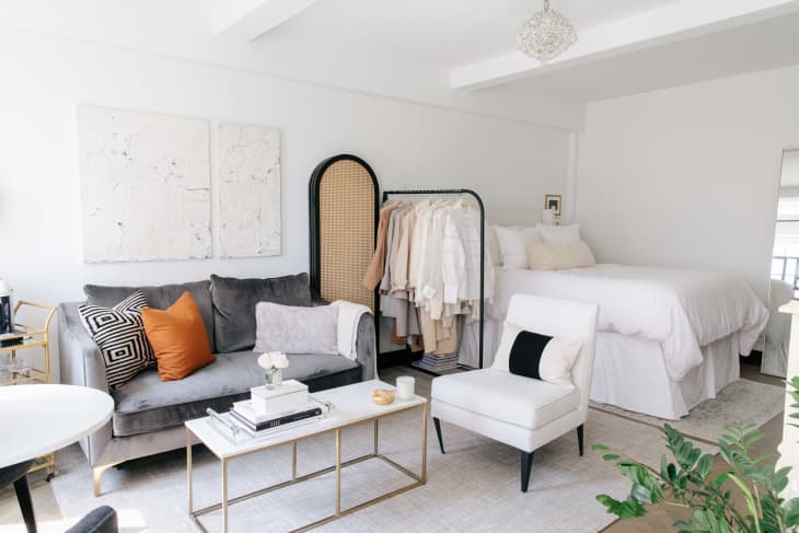 Studio apartment with a gray sofa, white armchair, bed, clothing rack, and modern decor.