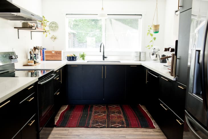Modern kitchen with black cabinets, white countertops, red patterned rug, and hanging plants by a large window.
