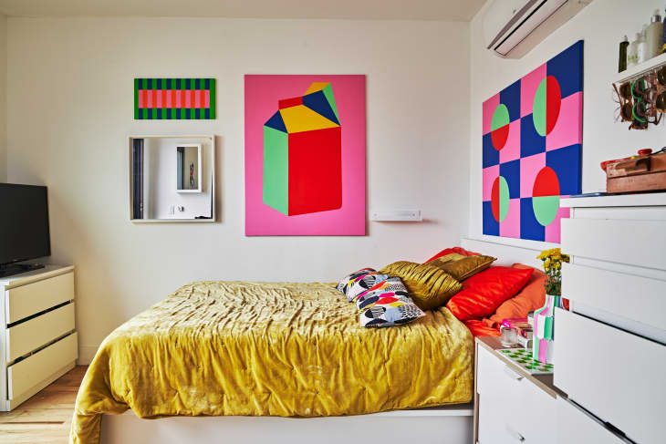 Bedroom with colorful abstract art, mustard bedspread, patterned pillows, and white furniture.