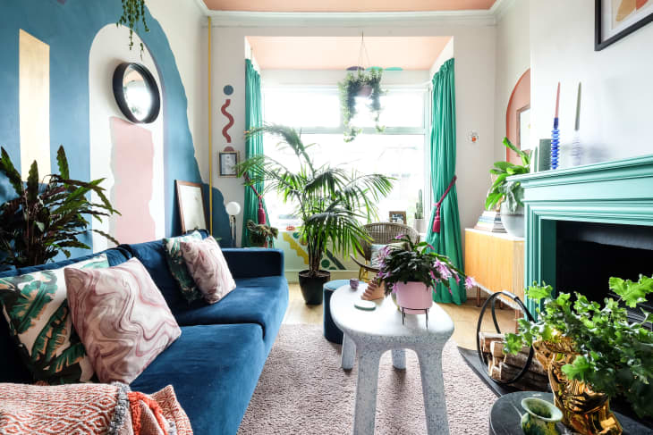Colorful living room with blue sofa, green curtains, plants, abstract wall art, and a mint green fireplace.