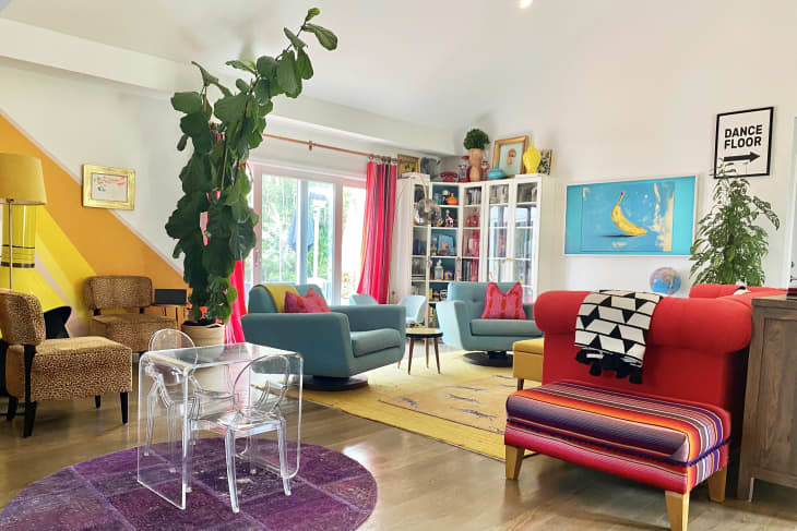 Colorful living room with blue sofas, red armchair, large plant, and vibrant decor, including a banana painting and glass table.