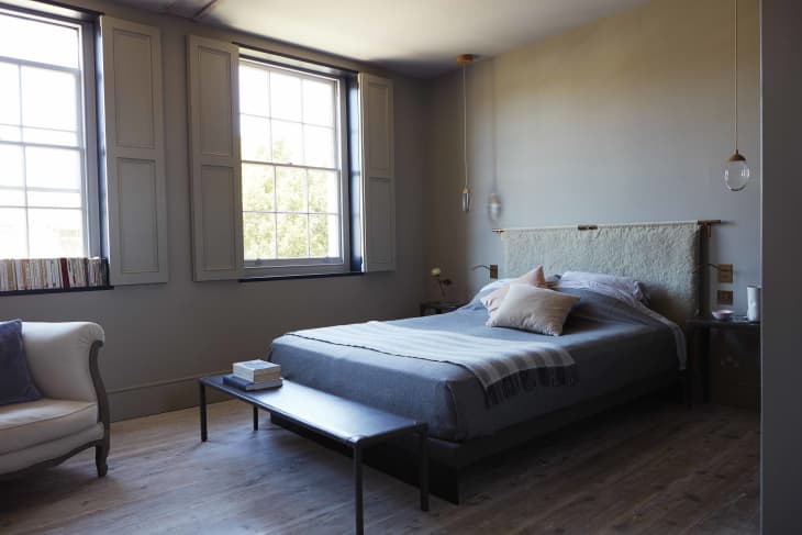 Bedroom with a gray bed, beige pillows, hanging lights, and two large windows. A small table holds books and decor.