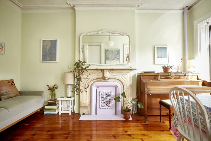Living room with a vintage fireplace, mirror, piano, and a dining table with a floral tablecloth.
