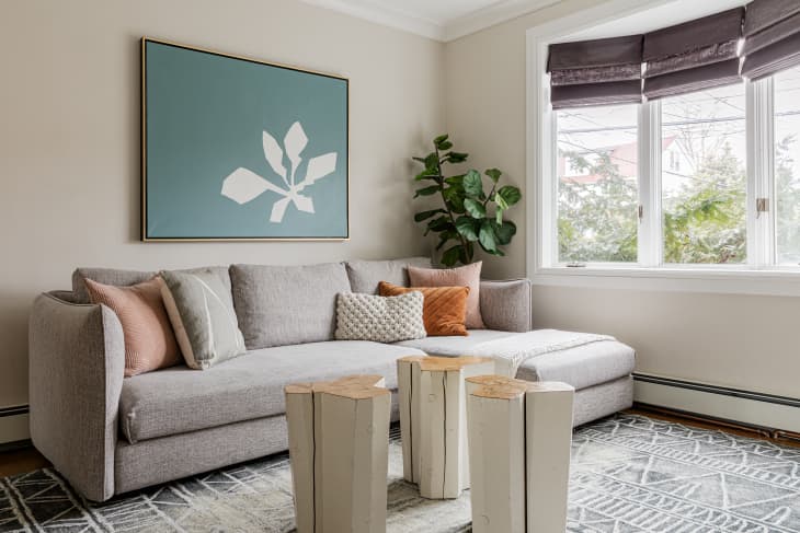 Gray sofa with assorted pillows, abstract wall art, wooden stump coffee tables, and a large plant by a window.