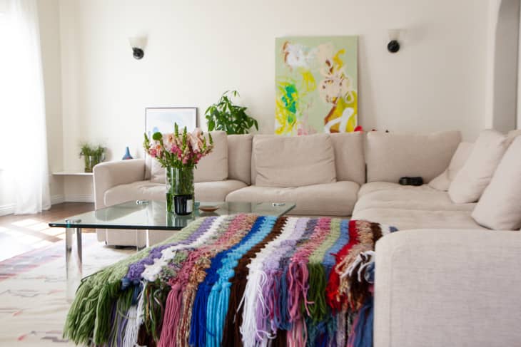 Cozy living room with white walls, tan sectional, and rainbow crocheted blanket at foot of sectional