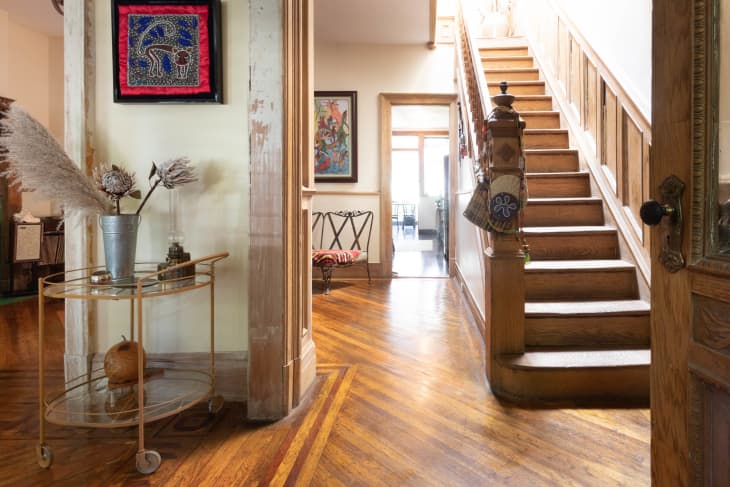 Wooden staircase with decorative hanging, art on walls, and a gold cart with dried flowers in a hallway.