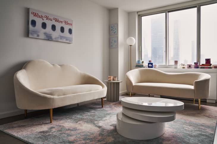 Two cream sofas in a living room with a round white coffee table, floor lamp, and a window view of skyscrapers.