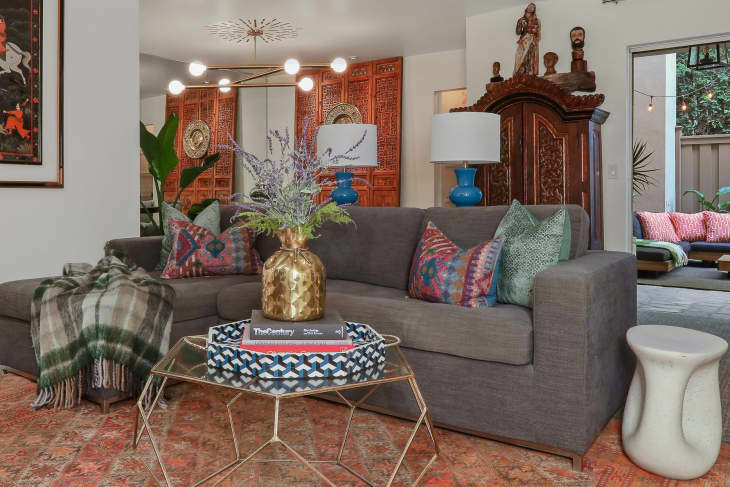 Gray sectional sofa with colorful pillows, gold vase, and blue lamps in a living room with ornate wooden decor and outdoor seating.