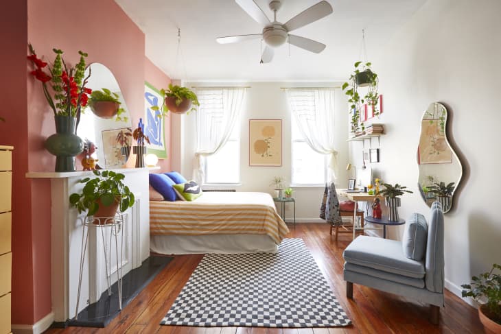 Bedroom with pink accent wall, striped bedding, plants, checkered rug, and a desk with a chair under a window.