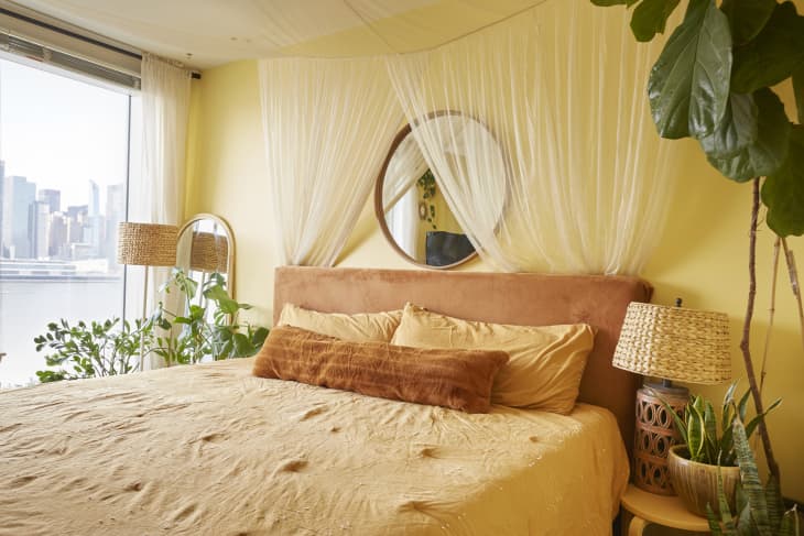 Cozy bedroom with a round mirror, peach headboard, yellow bedding, wicker lamps, and lush plants by a large window.
