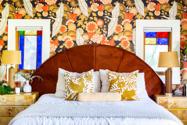 A bed with a brown headboard in front of busy patterned wallpaper and stained glass windows.