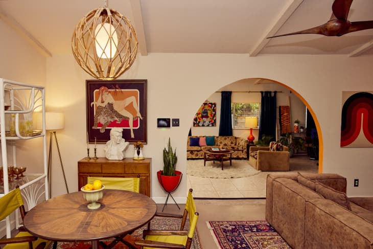 Mid-century modern living room with a round wooden table, yellow chairs, art pieces, and a large archway leading to a cozy seating area.
