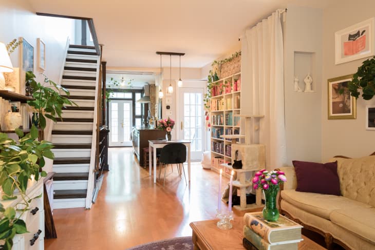 Cozy living room with beige sofa, bookshelves, plants, and a dining table with flowers under pendant lights.