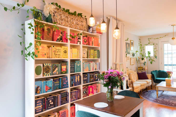 Cozy living room with bookshelves, a table with flowers, a cream sofa, and green armchairs, decorated with hanging plants.