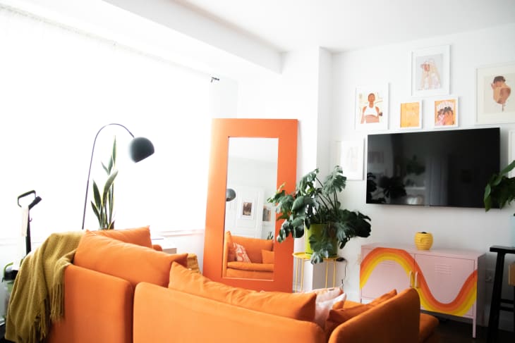 Bright living room featuring an orange sofa, large mirror, green plants, and colorful wall art above a TV.