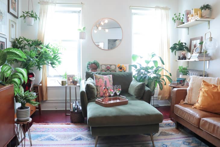 Cozy living room with green chaise, plants, round mirror, and decorative pillows, featuring a tray with glasses.