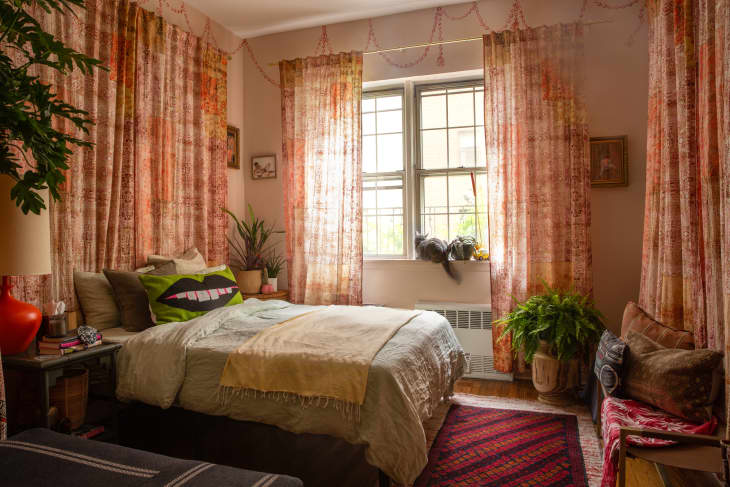 Cozy bedroom with patterned curtains, plants, a bed with colorful pillows, and a cat on the windowsill.