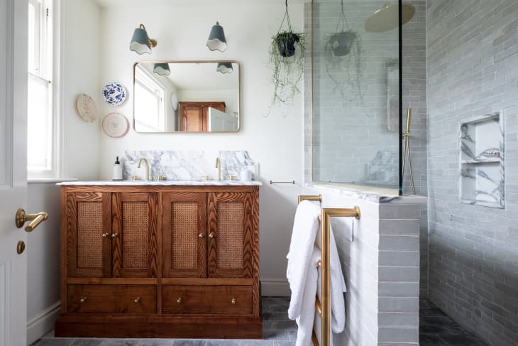 Two sconces above gold frame mirror hung above wooden vanity with marble top in newly renovated bathroom with a grey tiled bathroom.