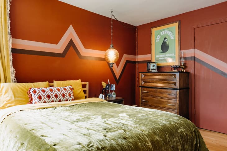 Zig-zag pink and brown decal runs along the wall of rust colored bedroom with neatly made bed with green velvet bedding.