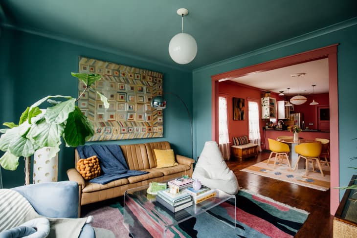 Quilt mounted above brown leather sofa in blue-green painted living room with globe pendant light and colorful rug on floor.