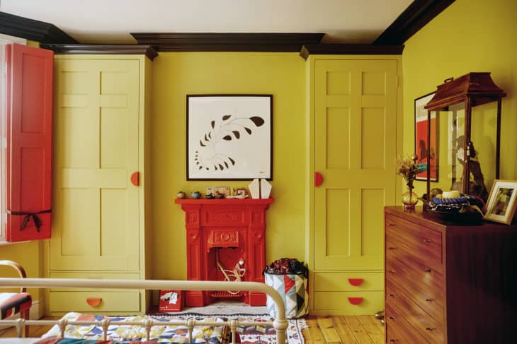built ins, mustard, red handles, half moon handles, cabinets, wardrobe, red shutters, wood floors, cream and multi colored rug, red mantle, multi colored chair with wood arm rest, wood dresser, cream metal foot board, black and white art, taxidermy bird in glass case