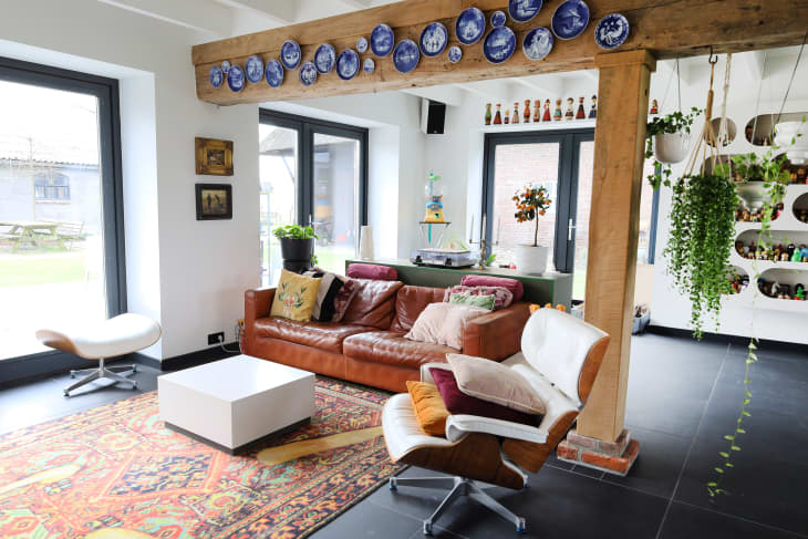 Living room with overhead wood beam decorated by blue and white plates, rust leather sofa, white cube coffee table, leather armchair with throw pillows, hanging plants, large windows, white walls