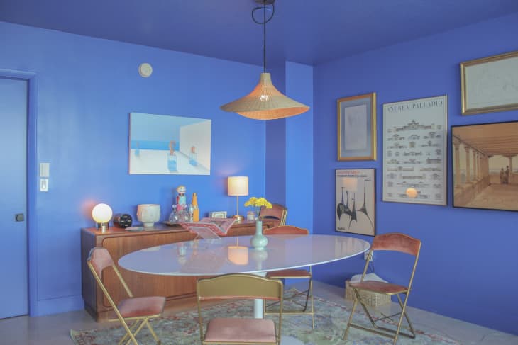 dining room with blue walls, white oval dining table, gold and pink velvet folding chairs, framed art, rattan/straw pendant light, wood credenza, patterned area rug.
