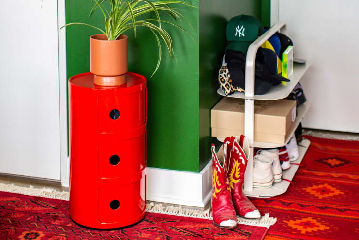 corner of bedroom with red patterned rug, shoe rack with hats and shoes, red small round drawer cabinet with plant, red cowboy boots on floor, green walls