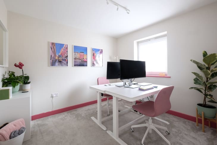 pink baseboards, beige carpet, and 2 white desks facing each other with computer monitors on each and pink swivel rolling desk chairs, 3 pictures hanging on wall with architecture in pink and blues