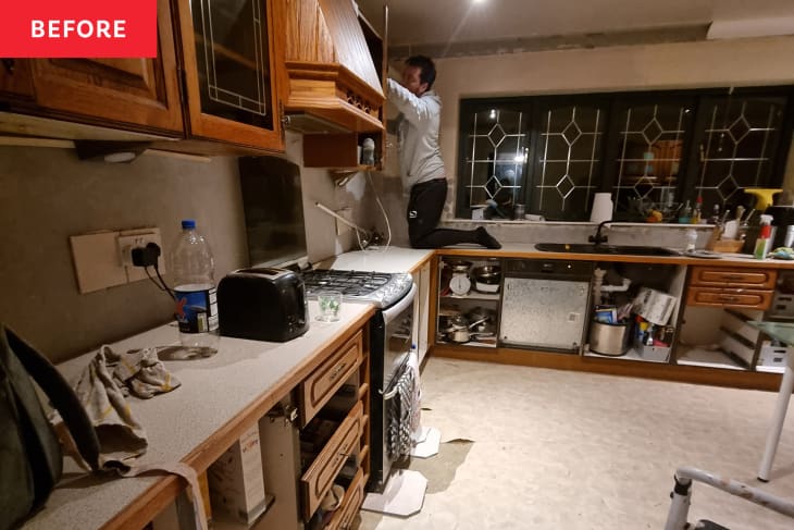 Cluttered kitchen with wooden cabinets, a man fixing a cabinet, and various items on the countertop.