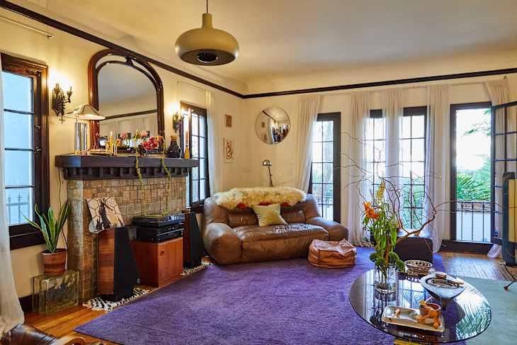 Cozy living room with a brown leather sofa, purple rug, fireplace, and large windows. Decor includes plants and a coffee table.