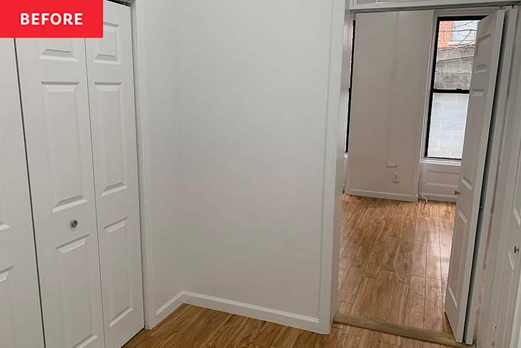 A small room with white walls, two closet doors, and light wooden flooring, leading to another room with windows.