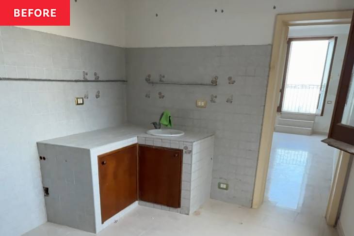 A small, unfinished kitchen area with gray tiled walls, a sink, and wooden cabinet doors, leading to a bright room.