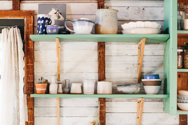 Rustic kitchen shelves with assorted pottery, bowls, and jars against a wooden wall.