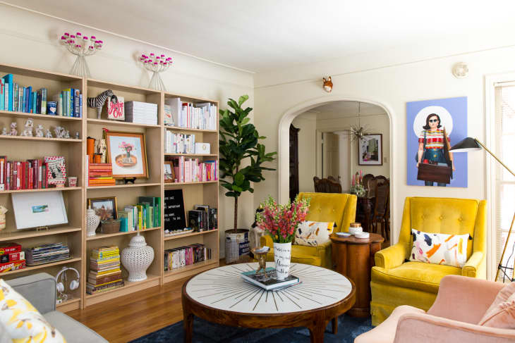 Living room with yellow armchairs, colorful bookshelves, a round coffee table, and a vibrant wall art piece.