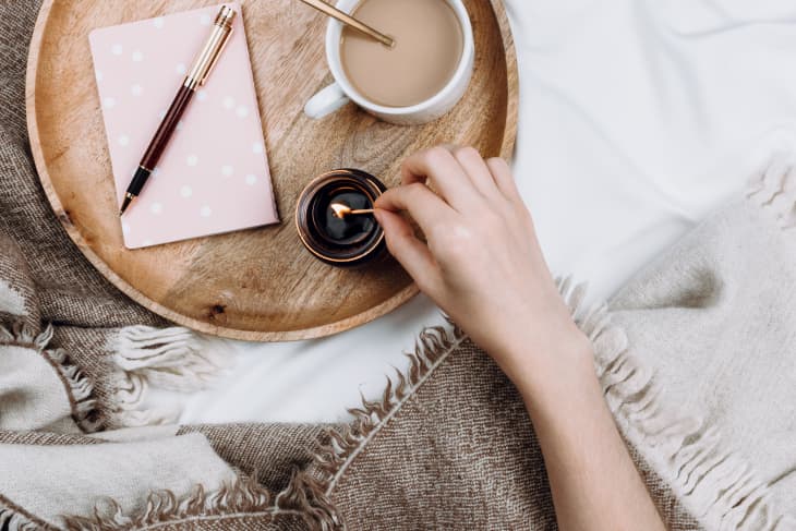 Cozy flatlay with wooden tray, cup of coffee or cocoa, candle, notebooks on white sheets and blankets