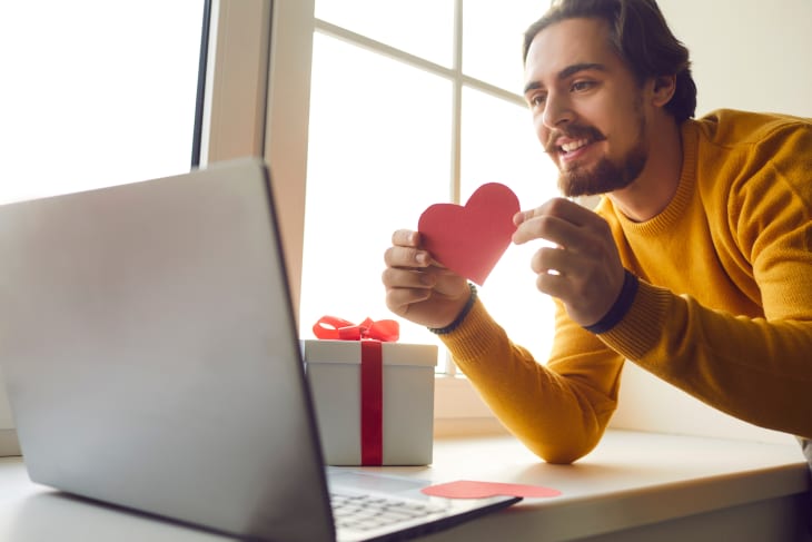 Closeup happy young man in love holding red heart-shaped card and showing it to girlfriend during video call on Saint Valentine's Day. Virtual date and long-distance relationship in quarantine concept
