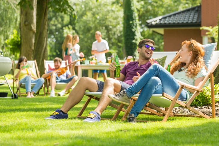 People relaxing on deck chairs in a sunny garden, holding drinks, with a barbecue and friends in the background.