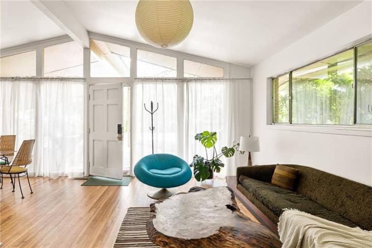 Mid-century modern living room with a blue chair, wooden coffee table, green sofa, and large windows with sheer curtains.