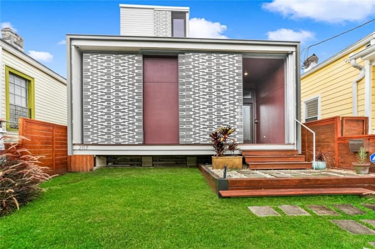 Modern house with geometric facade, wooden steps, and green lawn, flanked by red and yellow houses.