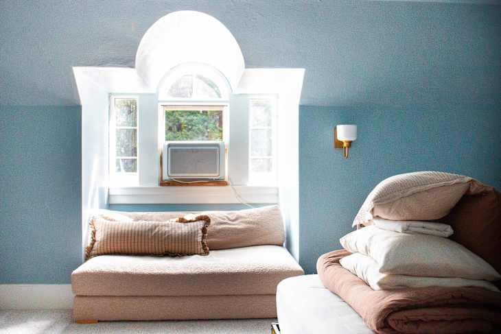 Cozy bedroom with a light blue wall, a window with an air conditioner, a beige couch, and stacked bedding.
