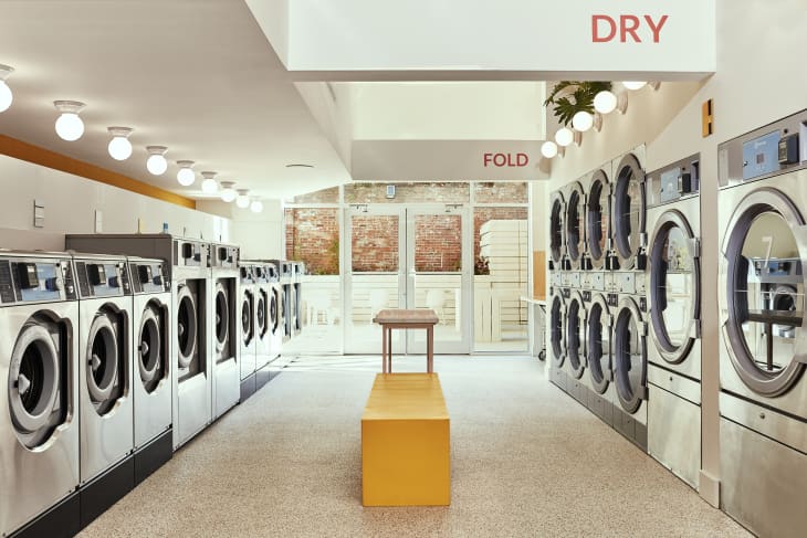 Modern laundromat with rows of stainless steel washers and dryers, yellow bench, and folding table.