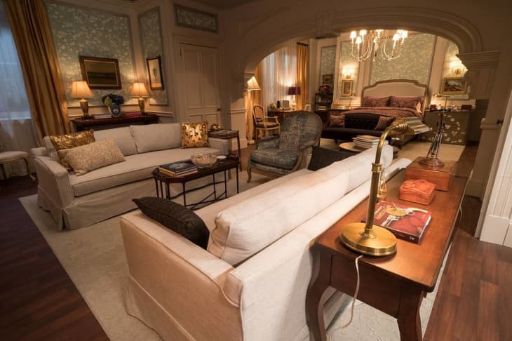 Elegant living room with cream sofas, gold accents, and a view into a bedroom with a chandelier and ornate bed.