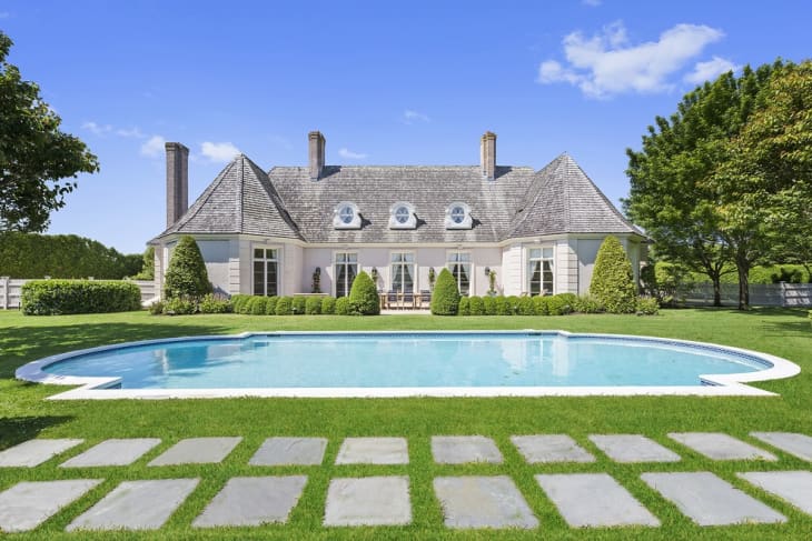 Elegant house with a shingled roof, surrounded by lush greenery, and a large oval pool in the foreground.