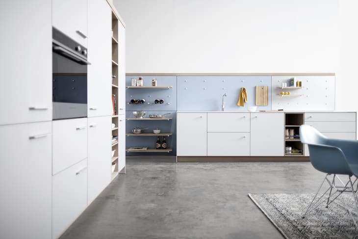 Modern kitchen with white cabinets, blue backsplash, open shelves, and a blue chair on a patterned rug.