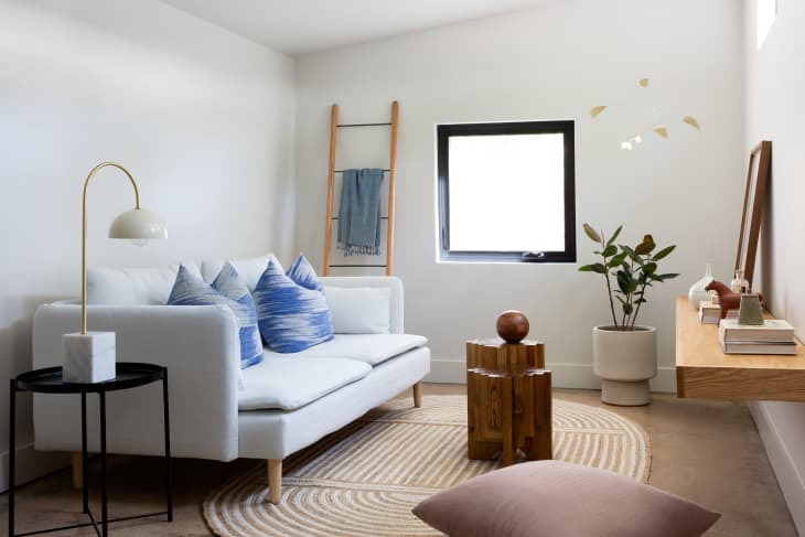 Minimalist living room with white sofa, blue pillows, wooden stool, potted plant, and a ladder shelf by the window.