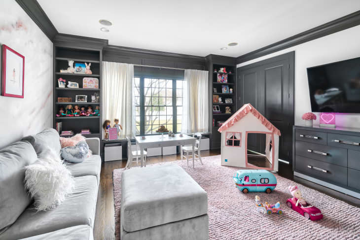 Playroom with gray sofa, toy house, dolls, and toy car on a pink rug, shelves with books and toys, and a wall-mounted TV.